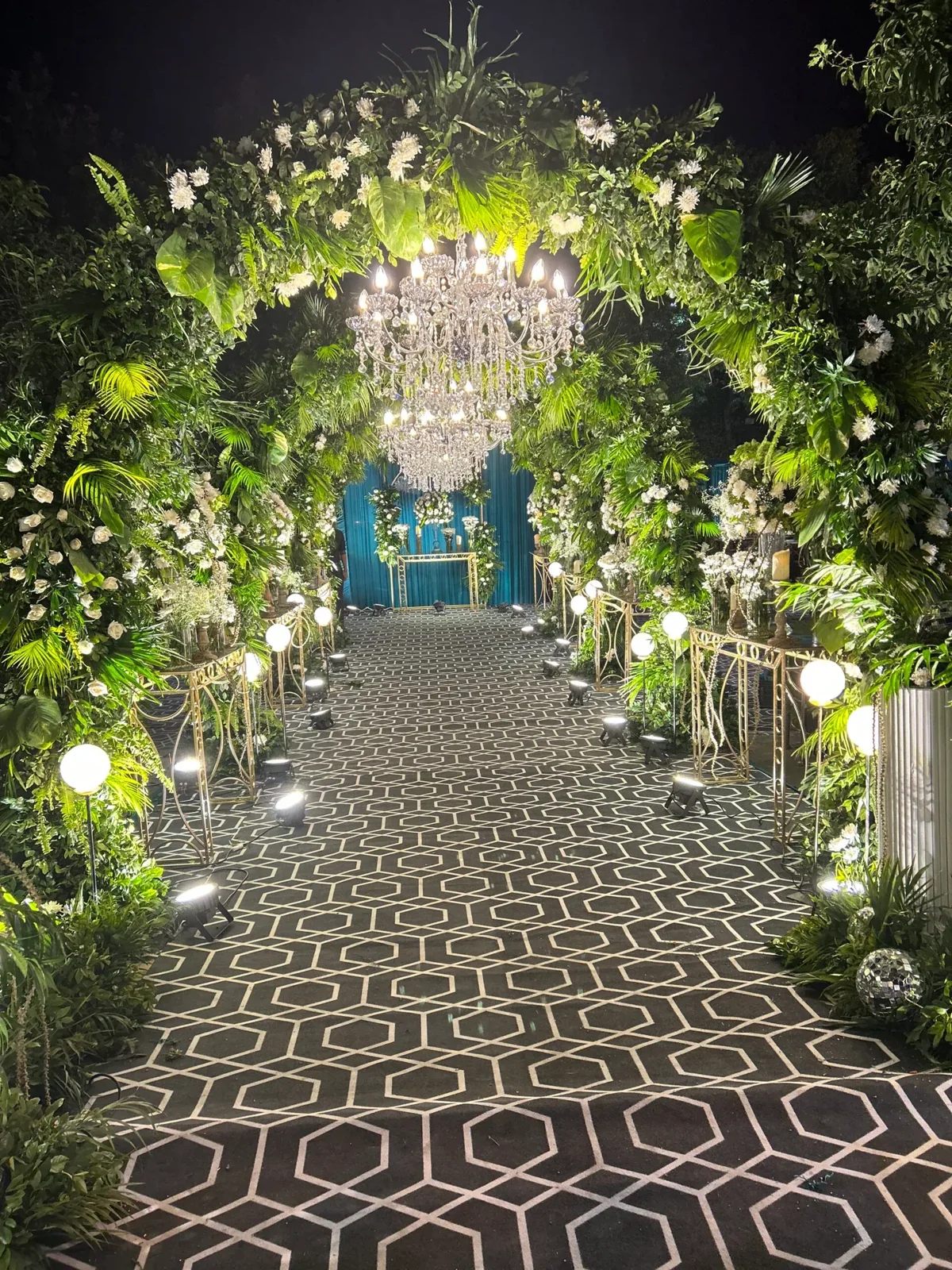 Lush green floral arches with crystal chandeliers above a geometric walkway